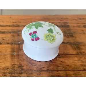 May include: A small, white, oval-shaped trinket box with a lid. The lid is decorated with green leaves, a pink and purple butterfly, and yellow-green clusters. The box sits on a wooden surface.