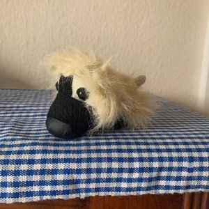 May include: A small, plush toy resembling a dog with white fur and a black face. The toy has black eyes and a black nose. The toy is lying on a blue and white checkered fabric.