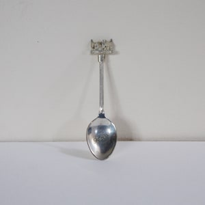May include: Silver souvenir spoon with a detailed design of a building on the handle. The spoon is on a white background.