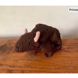 May include: A brown plush bison toy with pink horns and a dark brown nose, lying on a wooden surface. The toy has a soft, textured appearance. The bison toy is designed to resemble a bison.