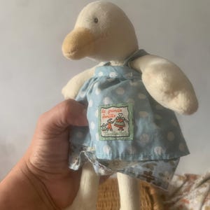 Pu&ograve; includere: Una bambola di anatra di peluche con corpo color crema, becco marrone e vestito blu a pois bianchi. L'abito ha una piccola toppa ricamata con testo e un disegno. Le zampe dell'anatra sono bianche.