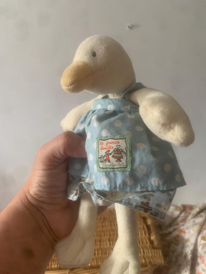 Pu&ograve; includere: Una bambola di anatra di peluche con corpo color crema, becco marrone e vestito blu a pois bianchi. L'abito ha una piccola toppa ricamata con testo e un disegno. Le zampe dell'anatra sono bianche.