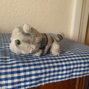 May include: A gray and white striped plush cat toy with blue and gold accents. The cat is lying on a blue and white checkered fabric.