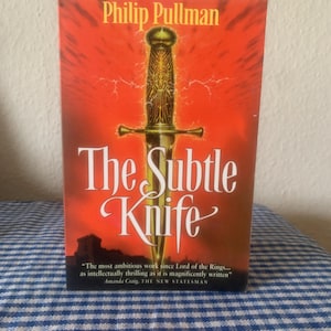 Peut inclure: Couverture du livre "The Subtle Knife" de Philip Pullman. Elle présente un poignard doré sur un fond orange vif. Le titre est en blanc, avec une citation d'Amanda Craig. Le nom de l'auteur est en haut.
