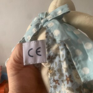 Pu&ograve; includere: Una bambola di coniglio di peluche con un vestito azzurro a pois bianchi e una gonna con stampa floreale. La bambola ha un'etichetta con le lettere "CE".