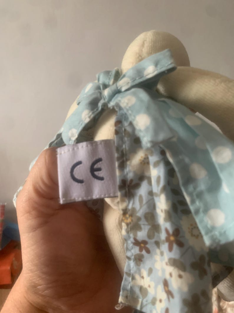 Pu&ograve; includere: Una bambola di coniglio di peluche con un vestito azzurro a pois bianchi e una gonna con stampa floreale. La bambola ha un'etichetta con le lettere "CE".