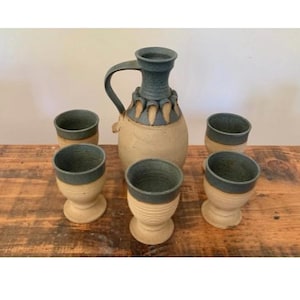Puede incluir: Un juego de jarra y tazas de cerámica hechas a mano. La jarra tiene una parte superior y un asa de color azul oscuro, con un cuerpo beige. Las tazas son de color beige con bordes azul oscuro. El juego se exhibe sobre una superficie de madera.