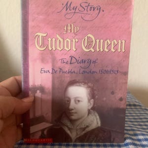 May include: A paperback book titled "My Tudor Queen" with a portrait of a woman on the cover. The book has a pink background with the title in ornate lettering. The author is Eva De Puebla, and the book is set in London from 1501-1513.