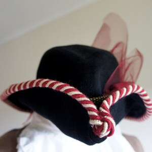 May include: A black felt hat with a wide brim and a red and white striped band. The hat has a sheer red mesh accent on the side.
