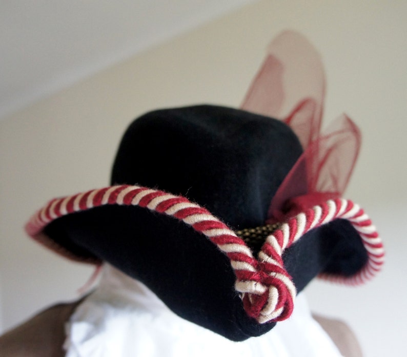 May include: A black felt hat with a wide brim and a red and white striped band. The hat has a sheer red mesh accent on the side.