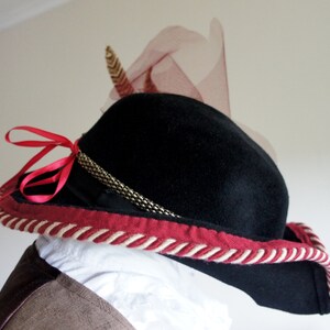 May include: A black felt hat with a wide brim, red and white trim, and a gold chain detail. A pink feather is attached to the top of the hat.