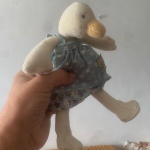 Pu&ograve; includere: Una bambola di anatra di peluche con testa, becco e arti color crema. L'anatra indossa un vestito azzurro con pois bianchi e una gonna floreale. La bambola ha una piccola etichetta sul vestito.