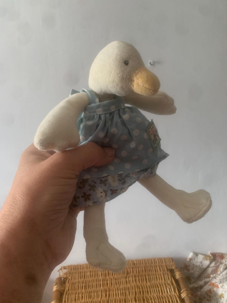 Pu&ograve; includere: Una bambola di anatra di peluche con testa, becco e arti color crema. L'anatra indossa un vestito azzurro con pois bianchi e una gonna floreale. La bambola ha una piccola etichetta sul vestito.