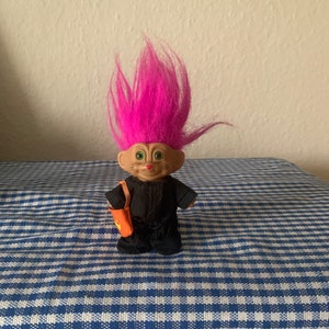 May include: A small pink-haired troll doll wearing a black jumpsuit and carrying an orange bag. The doll is standing on a blue and white checkered surface.