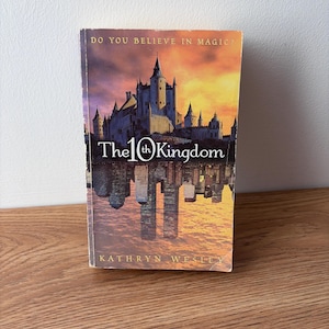 May include: A paperback book titled "The 10th Kingdom" with a castle illustration on the cover. The cover also features the text "Do You Believe in Magic?" and the author's name, Kathryn Wesley. The book is set on a wooden surface.