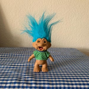May include: A blue-haired troll doll with a green and white polka dot shirt. The doll has a wide smile and is standing on a blue and white checkered surface.