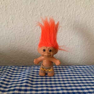 Peut inclure: Un petit troll en plastique avec des cheveux orange, un corps brun et un pagne jaune et brun. Le troll est debout sur une surface à carreaux bleus et blancs.