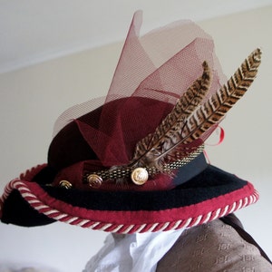 May include: A burgundy and black hat with a wide brim and a red feather trim. The hat has a burgundy tulle veil and a gold button accent.