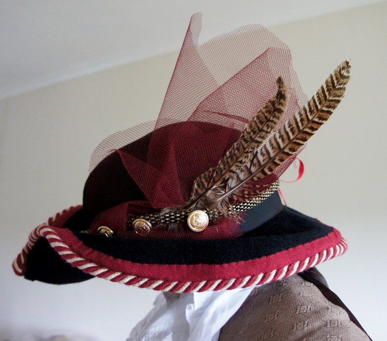 May include: A burgundy and black hat with a wide brim and a red feather trim. The hat has a burgundy tulle veil and a gold button accent.