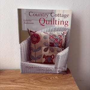 Peut inclure: Un livre à couverture souple intitulé Country Cottage Quilting par Lynette Anderson. La couverture présente un coussin décoratif beige avec des motifs appliqués floraux et d'arrosoir rouges et bleus, avec le texte 15 Quilt Projects Combining Stitchery with Patchwork.