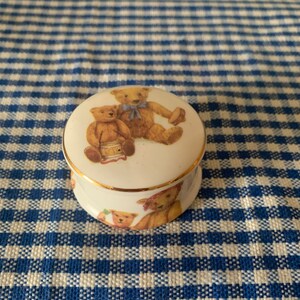 Könnte beinhalten: Eine weiße Keramik-Schmuckdose mit goldener Verzierung. Der Deckel zeigt ein Design mit drei Teddybären auf einem blau-weiß karierten Hintergrund.