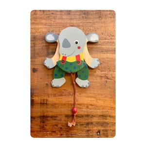 Op de afbeelding: Een houten trekkoord speelgoed in de vorm van een grijze neushoorn met een rode neus en roze wangen. De neushoorn draagt een geel shirt met rode bretels en een groene broek met stippen. Het speelgoed staat op een houten oppervlak.