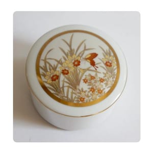 Könnte beinhalten: Eine runde, weiße Keramik-Schmuckdose mit goldenem Rand und Deckel mit Blumen- und Schmetterlingsdesign. Das Design umfasst gelbe Blumen, braune Blätter und einen braunen Schmetterling. Die Dose wird wahrscheinlich zur Aufbewahrung kleiner Gegenstände verwendet.