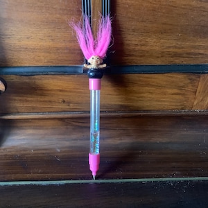 Pode incluir: Uma caneta de plástico transparente com um boneco troll rosa no topo. A caneta tem glitter dentro e uma tampa rosa.