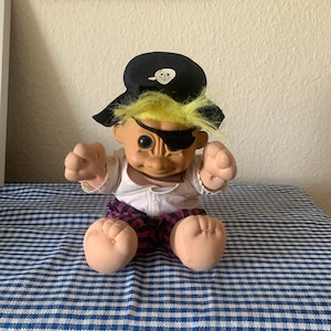 Puede incluir: Un muñeco de troll de peluche que lleva un sombrero de pirata negro con un diseño de calavera y huesos cruzados blancos. El troll tiene el pelo amarillo, una camisa blanca, pantalones rosa y negros y un parche en el ojo.