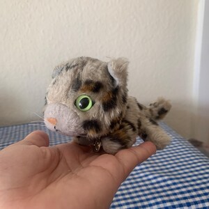 May include: A small, plush toy cat with brown and gray fur, green eyes, and a black collar. The cat is lying on its side with its head turned to the left.