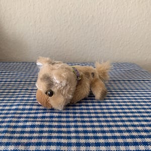 May include: A small, plush toy dog in shades of tan and brown, with a black eye and a small tail. The dog is wearing a purple collar. It is lying on a blue and white checkered surface.