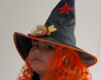 May include: A young person wearing a black witch hat with a red star and orange curly hair. The hat has a black brim and orange trim. The hat is decorated with a red star and a brown and yellow flower.