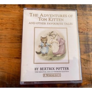 May include: A vintage cassette tape featuring a cover illustration of a girl and a cat from the book "The Adventures of Tom Kitten and Other Favourite Tales" by Beatrix Potter. The text on the cover reads "The Original and Authorized Edition" and "F. Warne & Co."