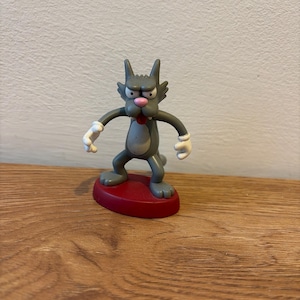 May include: A gray cartoon cat figurine with a red base. The cat has a grumpy expression, with a pink tongue sticking out. Its arms are outstretched, and it has white gloves. The figurine is standing on a red oval base.