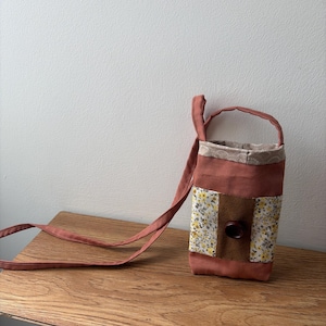 May include: A small, rectangular fabric pouch with a long shoulder strap in a muted terracotta color. The pouch features a central panel with a yellow and white floral pattern, a vertical brown fabric strip, and a large, dark brown circular button.