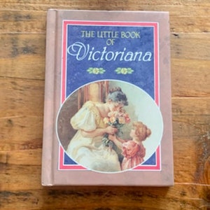 Könnte beinhalten: Ein Hardcover-Buch mit dem Titel "The Little Book of Victoriana" mit einem rosa Einband und einer blau-weißen Illustration auf der Vorderseite. Die Illustration zeigt eine Frau und ein Kind, die Blumen austauschen. Das Buch liegt auf einer Holzoberfläche.