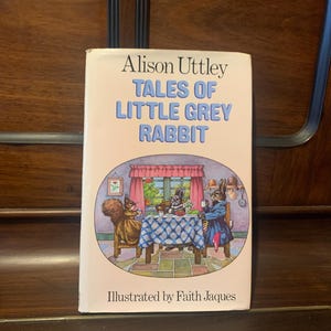Könnte beinhalten: Ein Buch mit dem Titel "Tales of Little Grey Rabbit" von Alison Uttley, illustriert von Faith Jaques. Der Umschlag zeigt eine farbenfrohe Illustration eines Eichhörnchens, eines Kaninchens und eines Dachses, die an einem Tisch mit einer karierten Tischdecke sitzen.