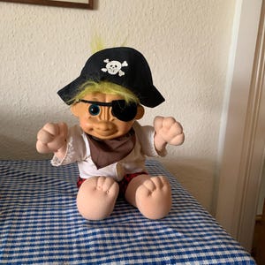 Puede incluir: Un muñeco de troll de peluche que lleva un sombrero de pirata negro con un diseño de calavera y huesos cruzados blancos. El troll tiene el pelo amarillo, un pañuelo marrón y una camisa blanca. El troll está sentado sobre una superficie a cuadros azules y blancos.