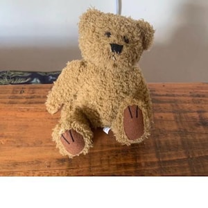 Puede incluir: Un oso de peluche marrón con ojos negros y una nariz marrón. El oso está sentado sobre una superficie de madera.