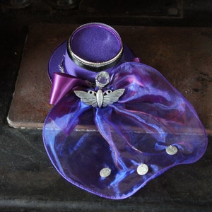 May include: A small purple top hat with a silver butterfly embellishment and a sheer purple veil. The hat is decorated with silver accents and a clear gem.