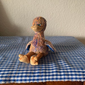 May include: A small, plush toy in the shape of an ostrich. The toy is brown and purple with blue eyes. It is sitting on a blue and white checkered surface.