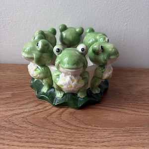 Op de afbeelding: Een keramische kandelaar met kikker-thema. De houder heeft een ring van groene kikkers met witte bloemaccenten, rondom een centrale ruimte. De basis heeft de vorm van een groen lelieblad. De kikkers hebben grote ogen en staan in een speelse pose.