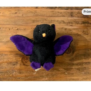 Peut inclure: Une peluche noire en forme de chauve-souris avec des ailes et des pieds violets, et un bec orange. La chauve-souris est posée sur une surface en bois. Les ailes sont déployées, et l'objet est un jouet.