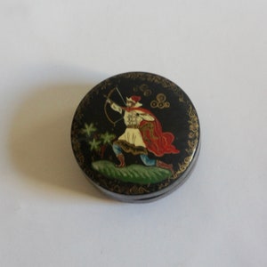 Op de afbeelding: Een ronde, zwart gelakte doos met een gouden rand en een geschilderd tafereel van een man in een rood-wit kostuum met een boog en pijl. De doos is versierd met gouden spiralen en stippen.
