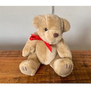 Puede incluir: Un oso de peluche marrón con un lazo rojo alrededor del cuello. El oso está sentado sobre una superficie de madera.