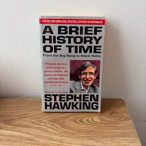 Peut inclure: Un livre de poche intitulé A Brief History of Time: From the Big Bang to Black Holes par Stephen Hawking. La couverture présente une photographie de Stephen Hawking et une citation du Sunday Times. Le texte est imprimé en noir et rouge.