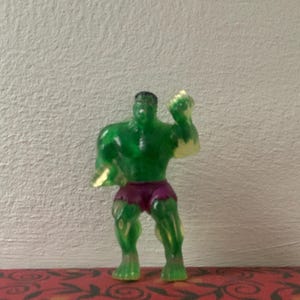 May include: A translucent green Hulk action figure with purple shorts. The figure is posed with one fist clenched and has black hair. The figure is standing on a red surface with a floral pattern.