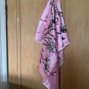 Può includere: Un foulard di seta rosa con una stampa floreale di fiori di magnolia bianchi e steli verdi. Il foulard è appeso a una porta di legno.