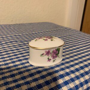 Peut inclure: Une petite boîte à bijoux en porcelaine de forme ovale avec un motif floral. La boîte est blanche avec une bordure dorée et présente des fleurs violettes et des feuilles vertes.