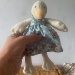 Pu&ograve; includere: Una bambola di peluche a forma di oca con corpo color crema, becco giallo e vestito blu a pois bianchi. L'abito presenta un motivo floreale sul fondo. La bambola ha le ali spiegate ed &egrave; alta circa 20 cm.